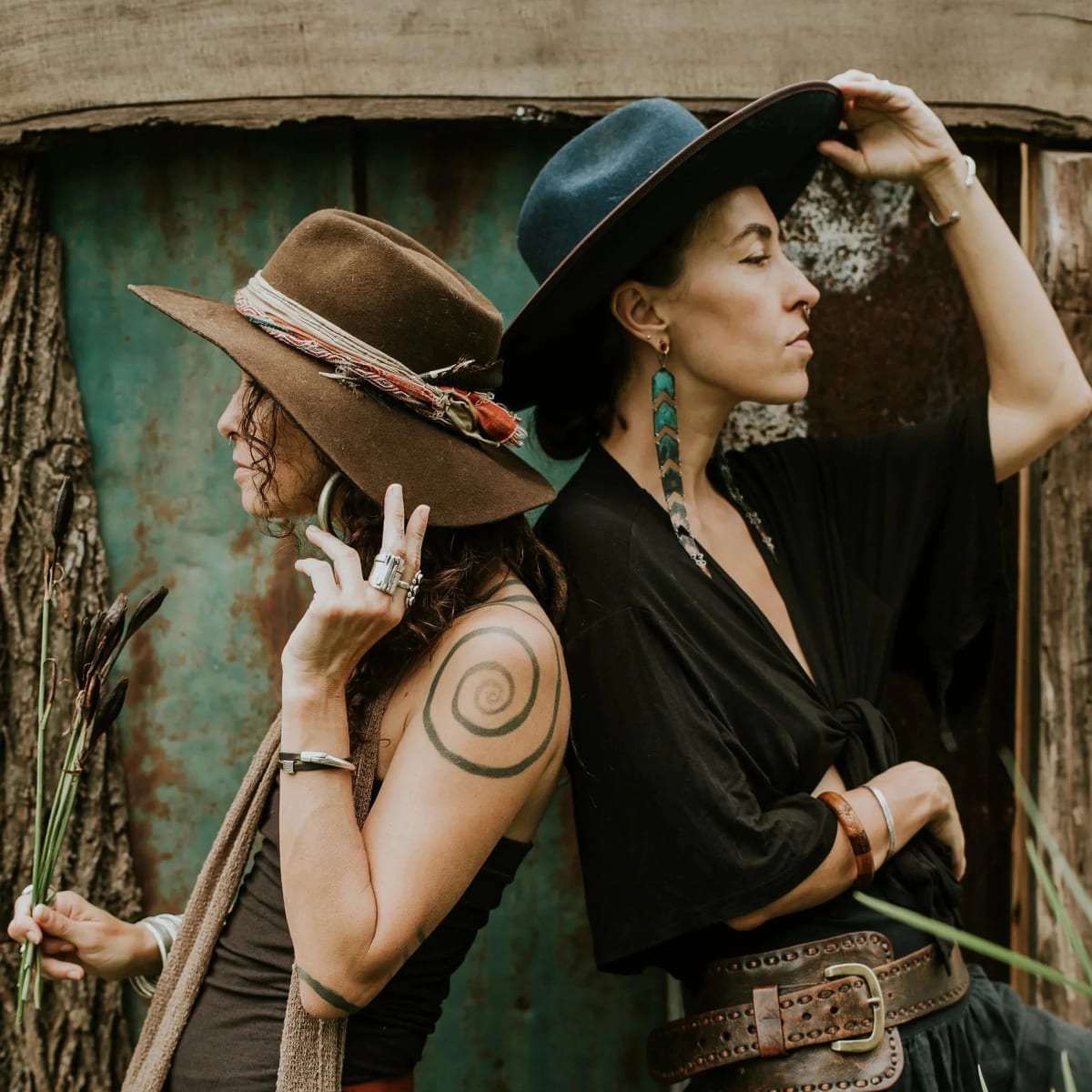 Leah Song and Chloe Smith of Rising Appalachia in wide-brimmed hats, standing back to back against a weathered teal wall